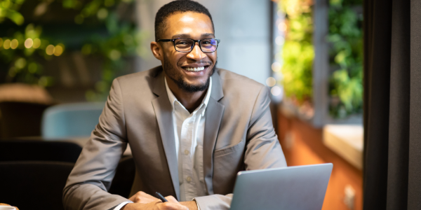 Smiling man in glasses and suit working on a laptop in an office with greenery in the background. The setting is professional and welcoming.